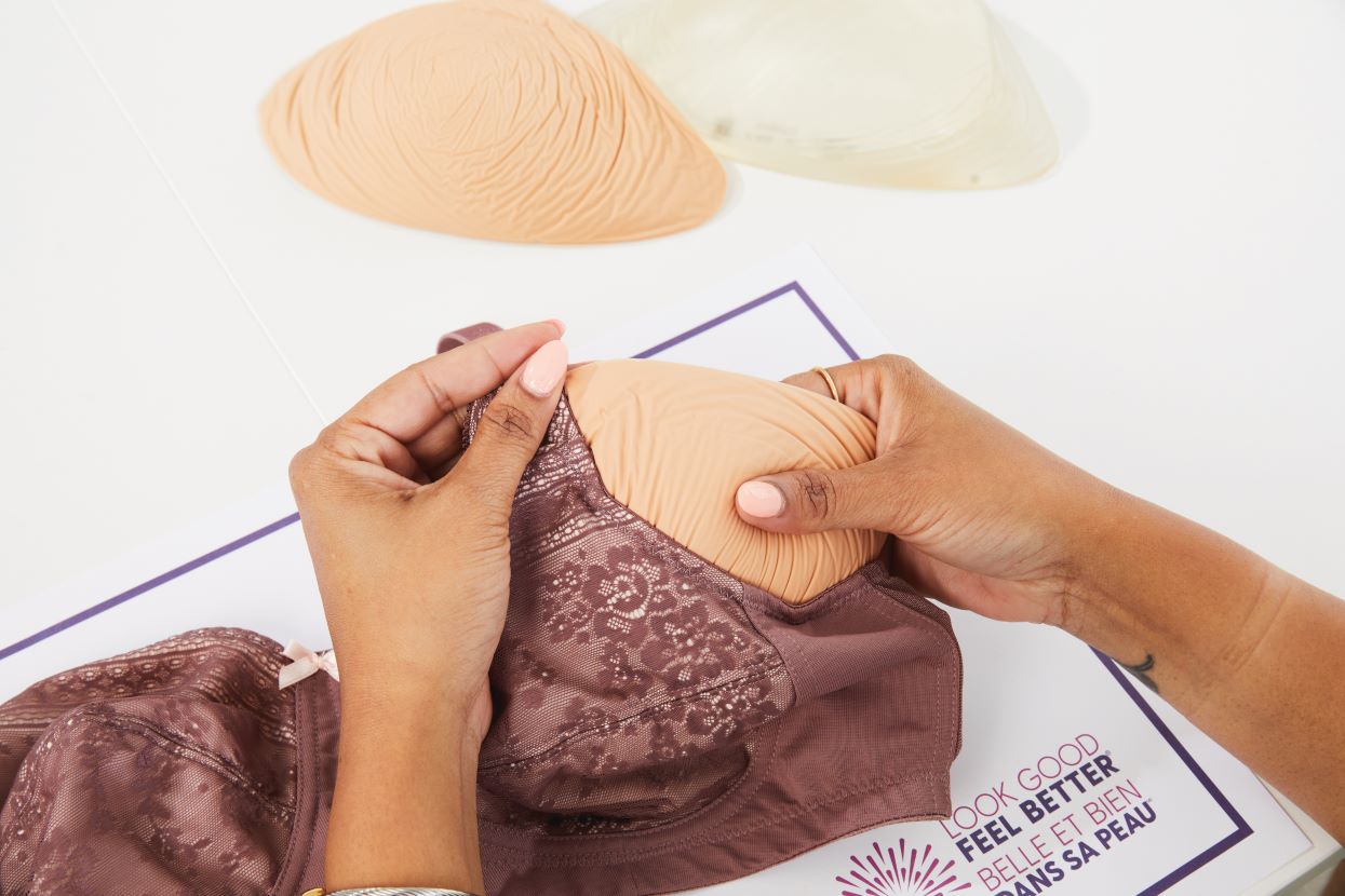 Hands placing a beige breast prosthesis into a mauve lace bra, with extra inserts and a Look Good Feel Better poster nearby.