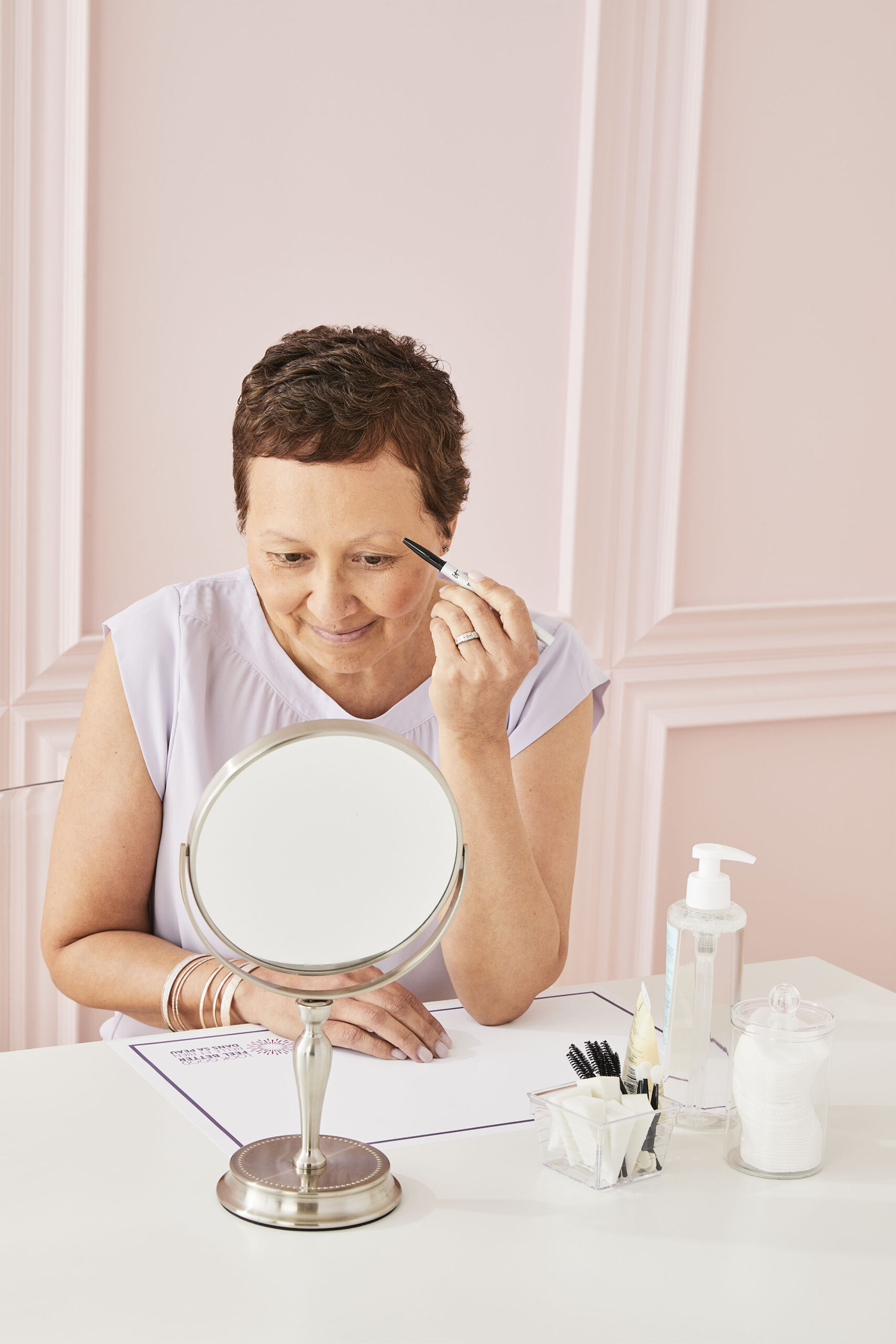 Smiling woman applies eyebrow makeup while looking into a small mirror on a table with beauty products.