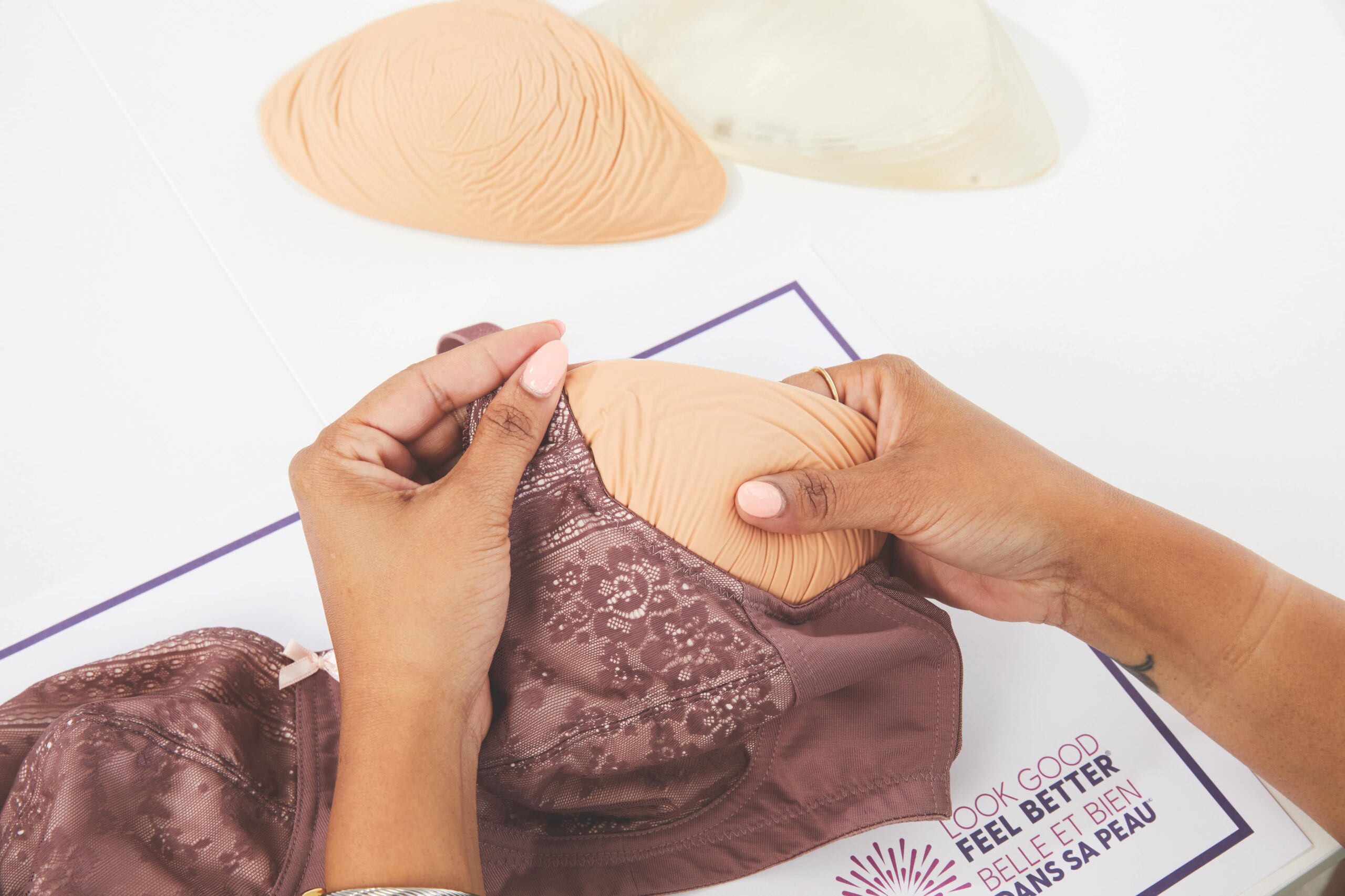 Des mains plaçant une prothèse mammaire beige dans un soutien-gorge en dentelle mauve, avec des inserts supplémentaires et une brochure Look Good Feel Better à proximité.