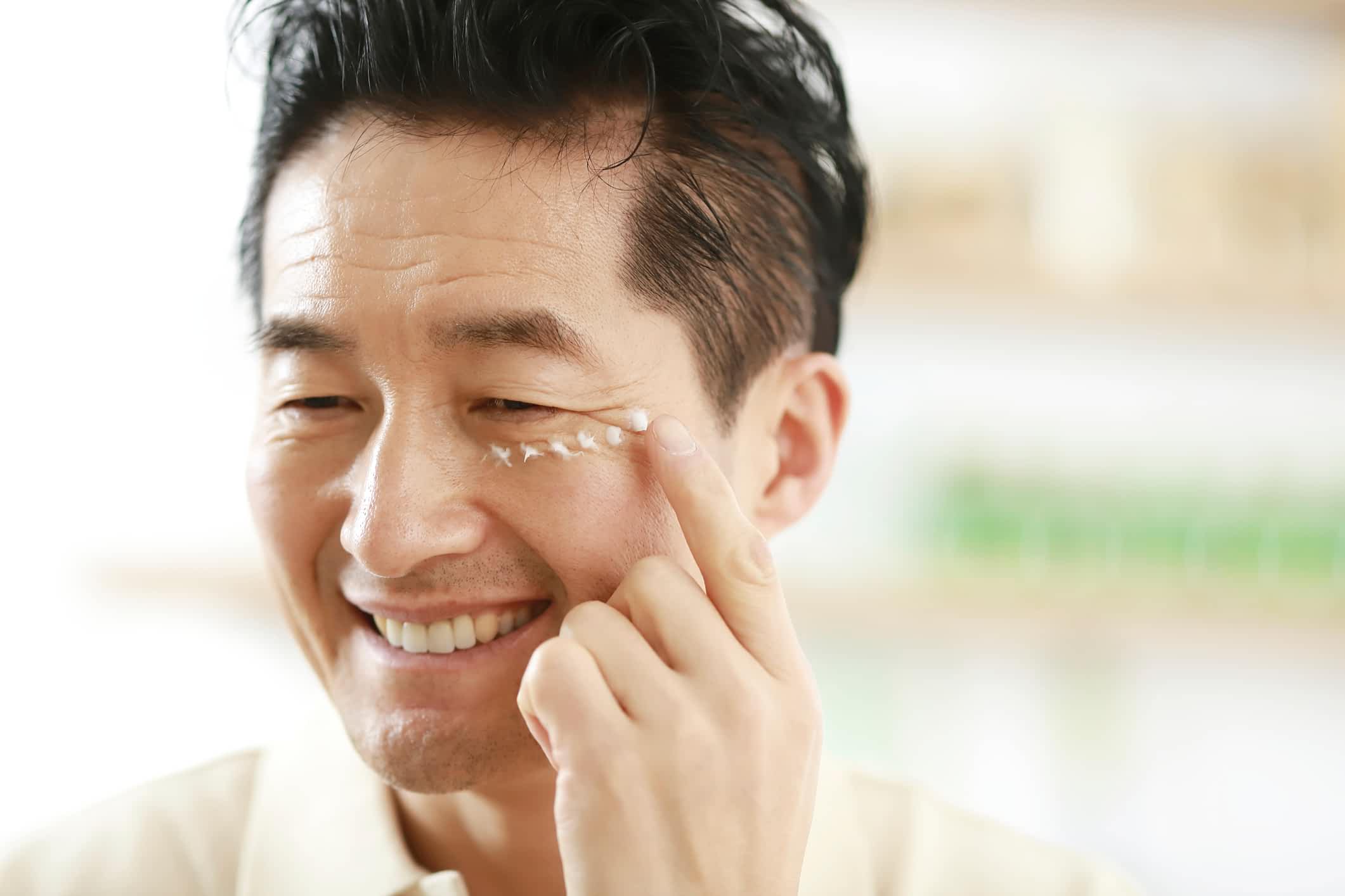 Smiling man applies eye cream under his right eye using his fingertip.
