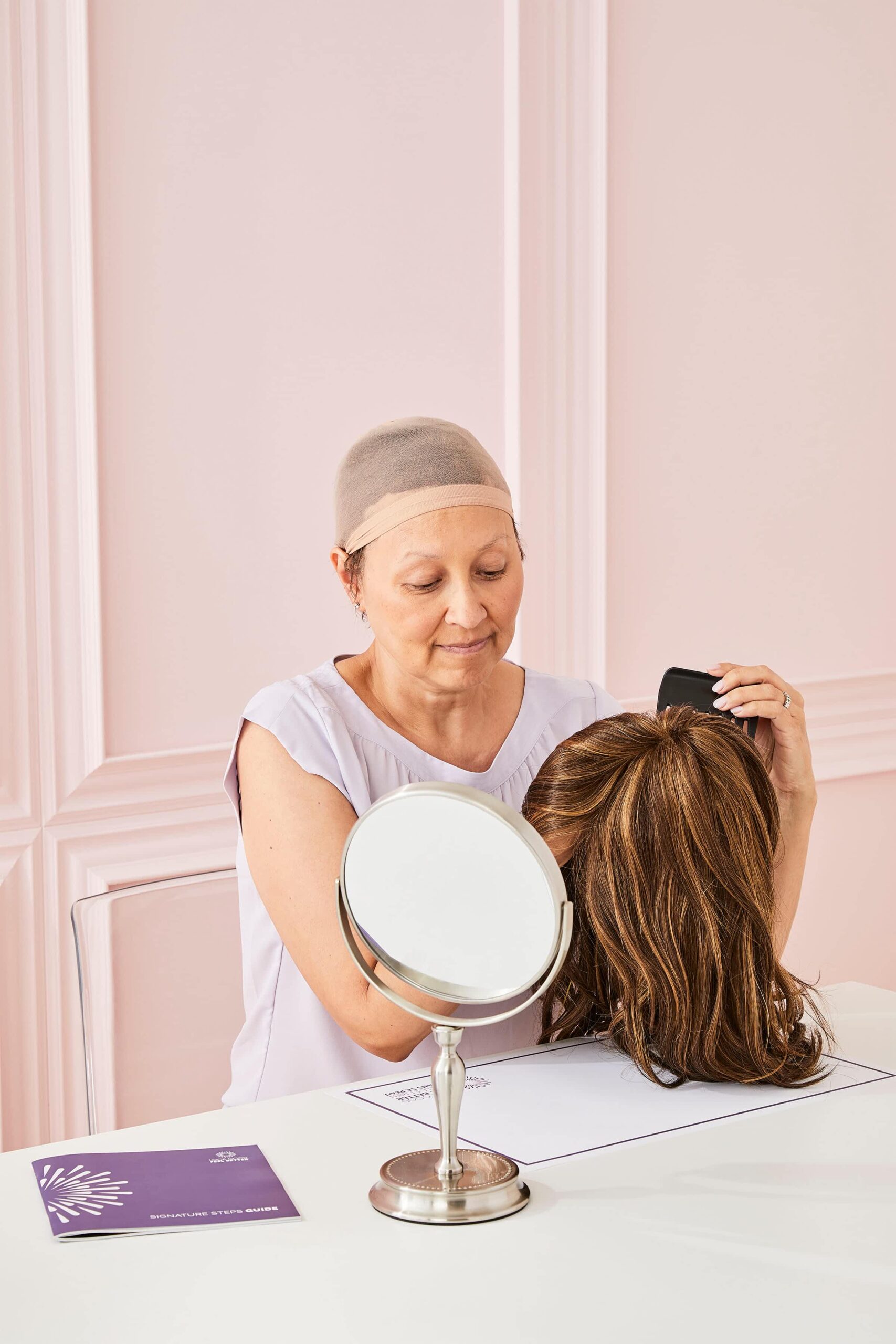 Woman wearing a wig cap sits at a table, holding a brown wig while looking at it in a round mirror, with a brochure beside her.