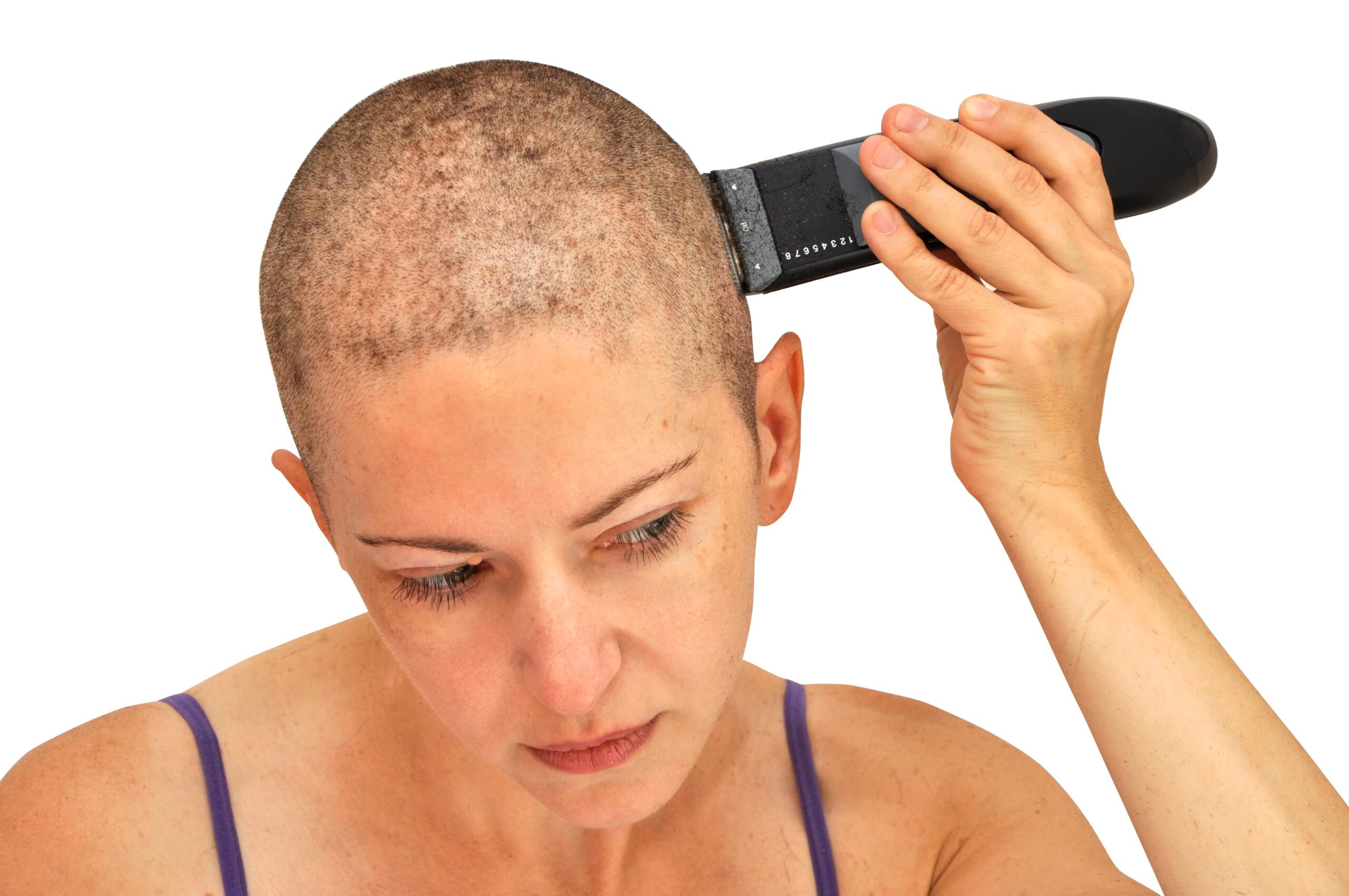 Woman with a shaved head uses clippers to shave remaining hair, wearing a thin strap top against a plain white background.