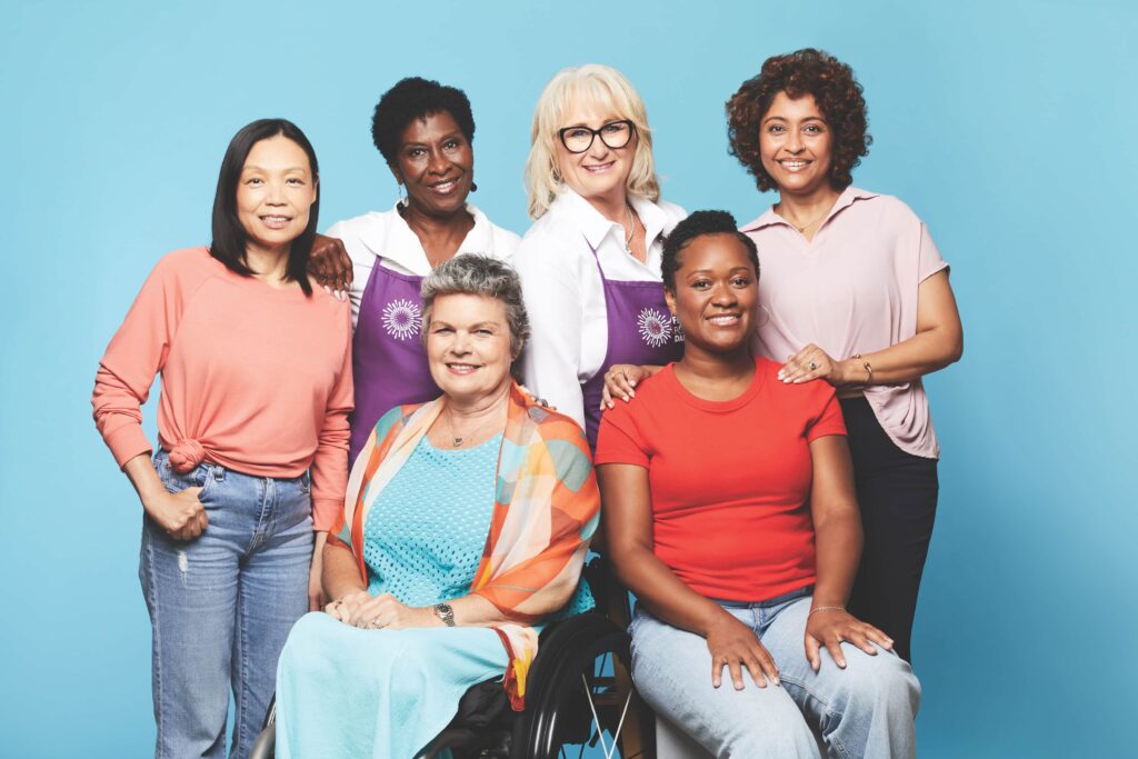 Two Look Good Feel Better volunteers stand while four workshop participants – two seated and two standing – pose together smiling in a bright room.