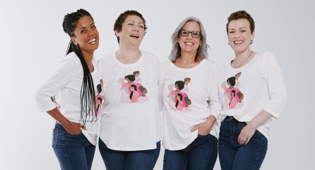 Quatre femmes souriant ensemble, vêtues de chemises blanches égales ornées de motifs roses, posant devant un fond uni.