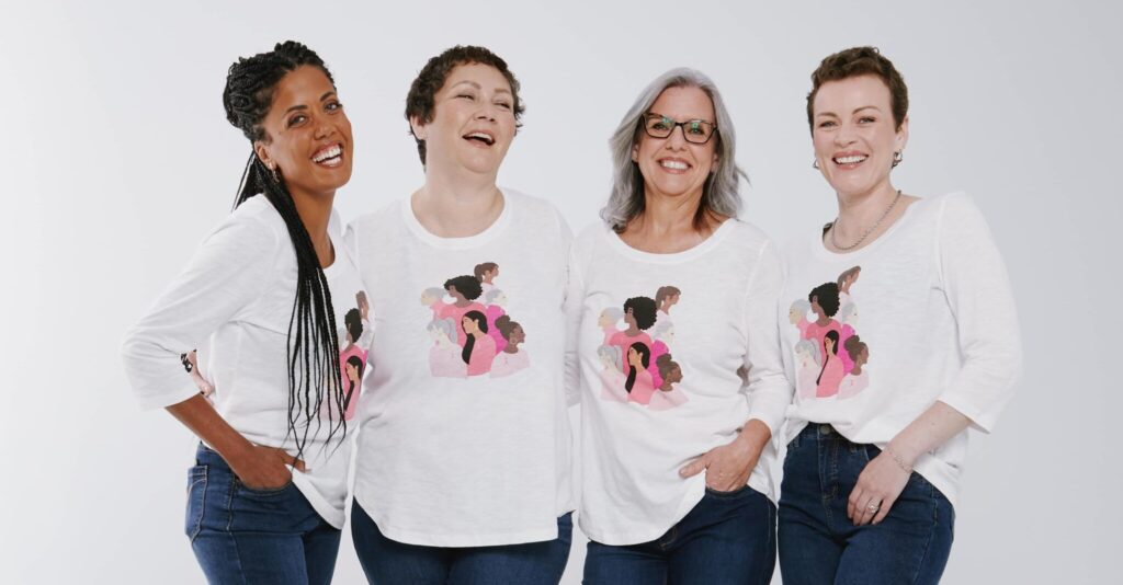 Four women smiling together in matching white shirts with pink designs, posing against a plain background.