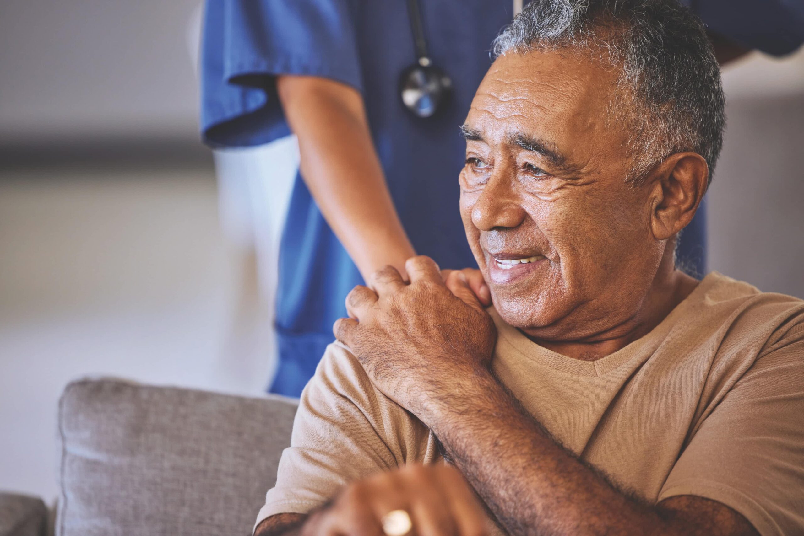 Smiling older man in a beige shirt sits on a couch as a healthcare worker in blue scrubs rests a supportive hand on his shoulder.