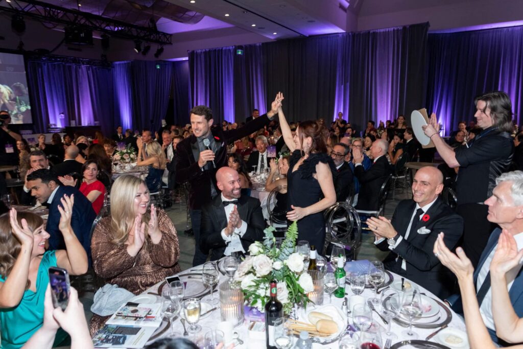 Guests seated at round dinner tables applaud as a woman is recognized and raises her hand with the host during the Mirror Ball gala, with elegant décor and stage lighting in the background.