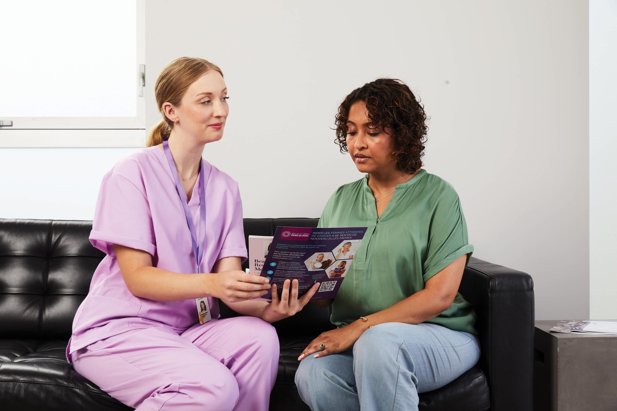 A nurse in light purple scrubs sitting next to a woman with a green blouse who is holding a brochure.