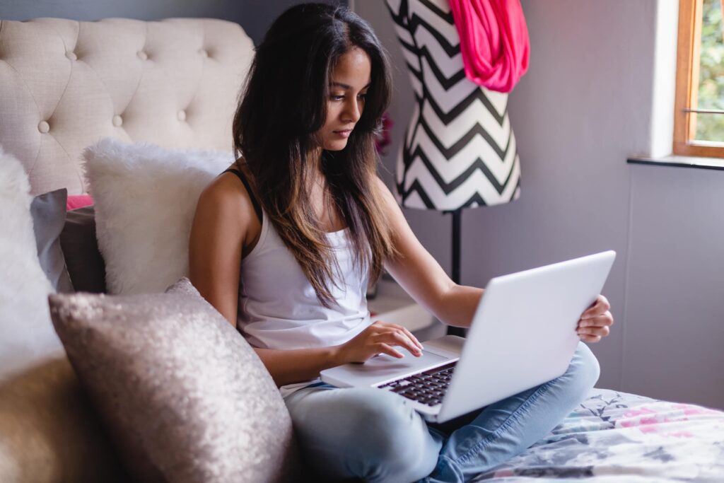 Une adolescente assise sur son lit devant un ordinateur portable, avec un mannequin de couture à motifs en arrière-plan.