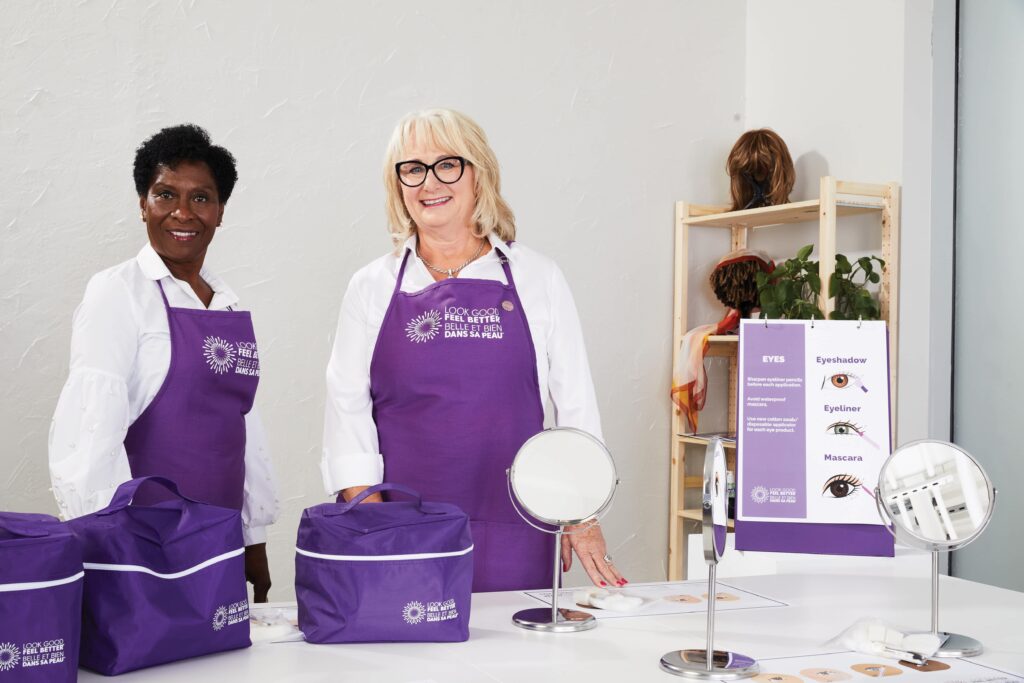 Two Look Good Feel Better volunteers stand in a workshop behind a table set with mirrors and cosmetic kits.