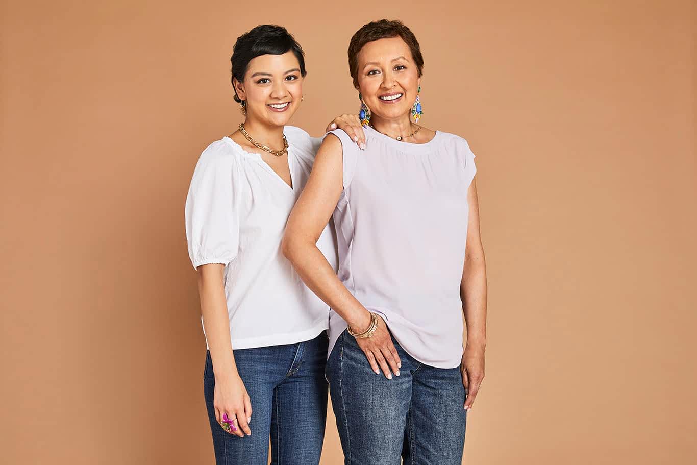 Two women with short hair smiling and posing together in white tops and jeans against a beige background.