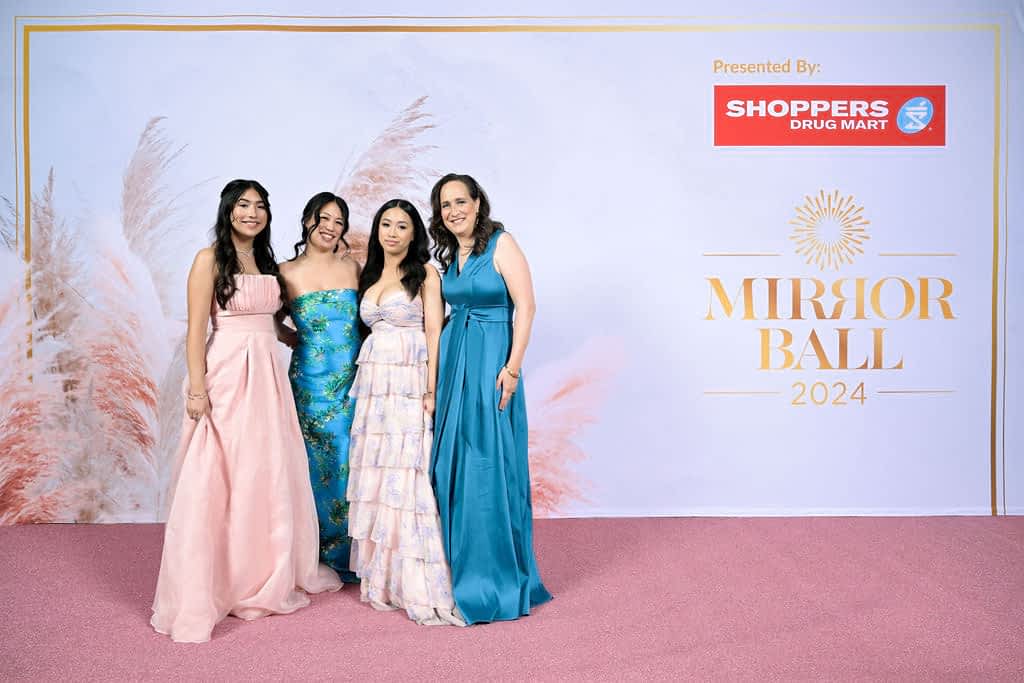 Four women in ballgowns posing in front of a Shopper's Drug Mart Mirror Ball background.