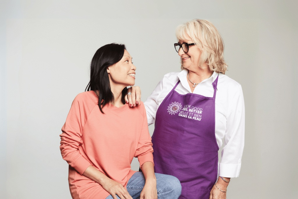 A smiling woman in a coral sweater sits while another woman in a purple "Look Good Feel Better" apron rests her hand on her shoulder.