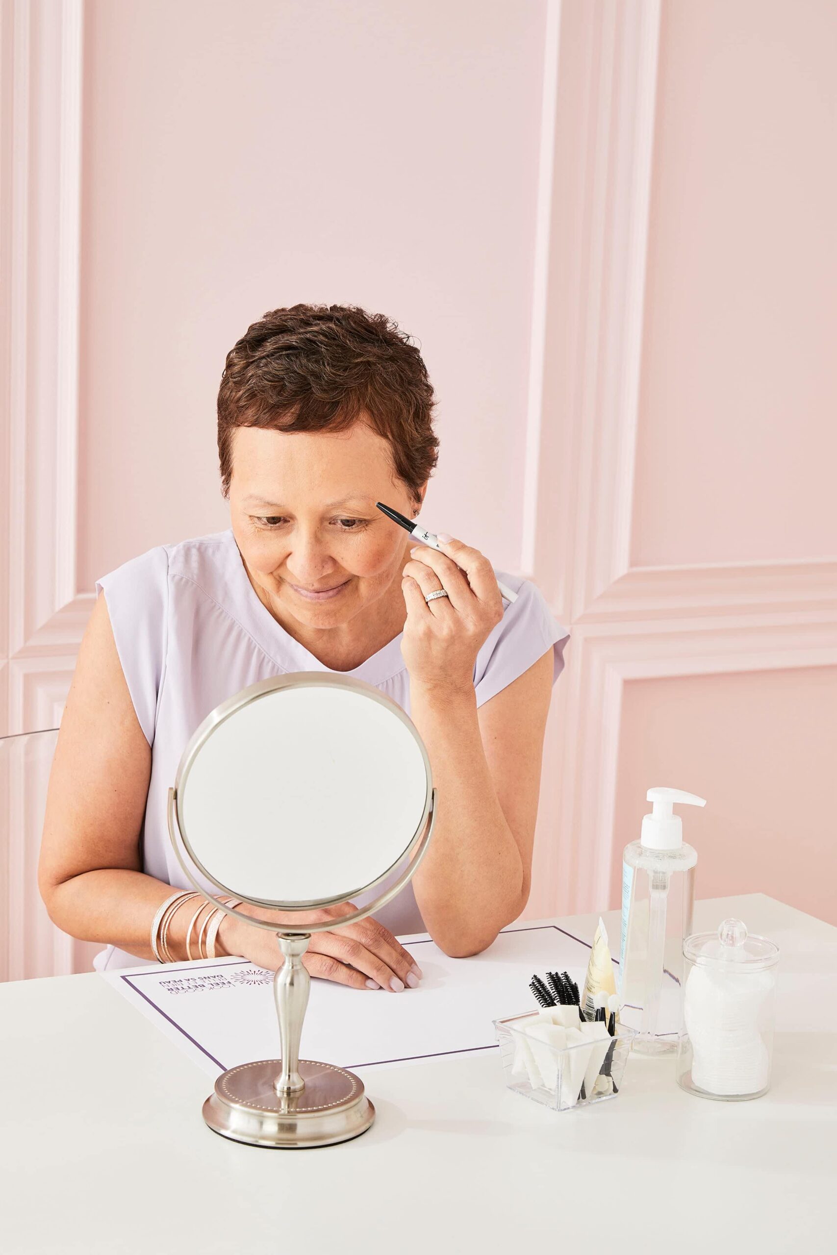 Smiling woman applies eyebrow makeup while looking into a small mirror on a table with beauty products.