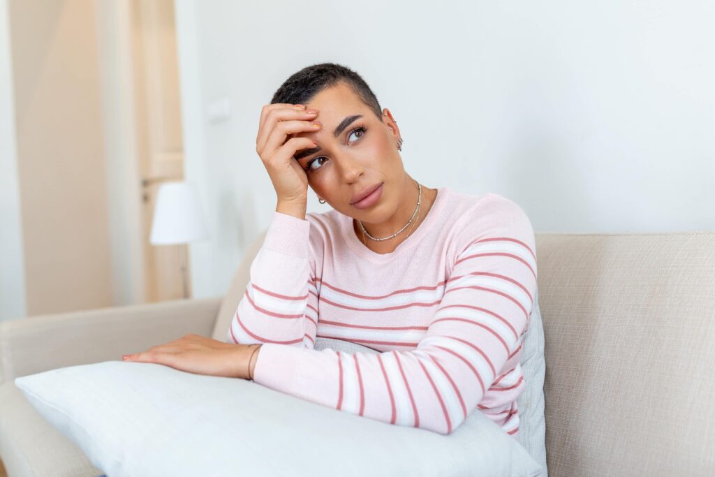 Femme réfléchie aux cheveux courts, appuyant sa tête sur sa main alors qu'elle est assise sur un canapé, l'air inquiet dans un cadre domestique calme.