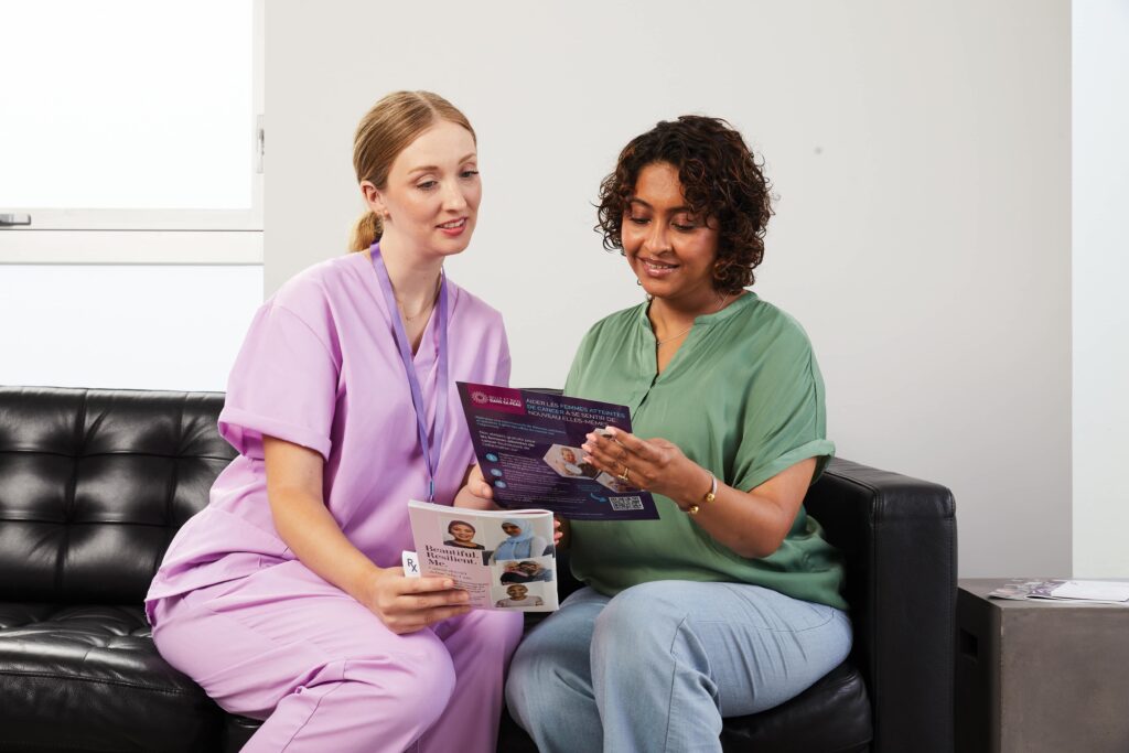 A nurse in light purple scrubs sitting next to a woman with a green blouse who is holding a brochure.