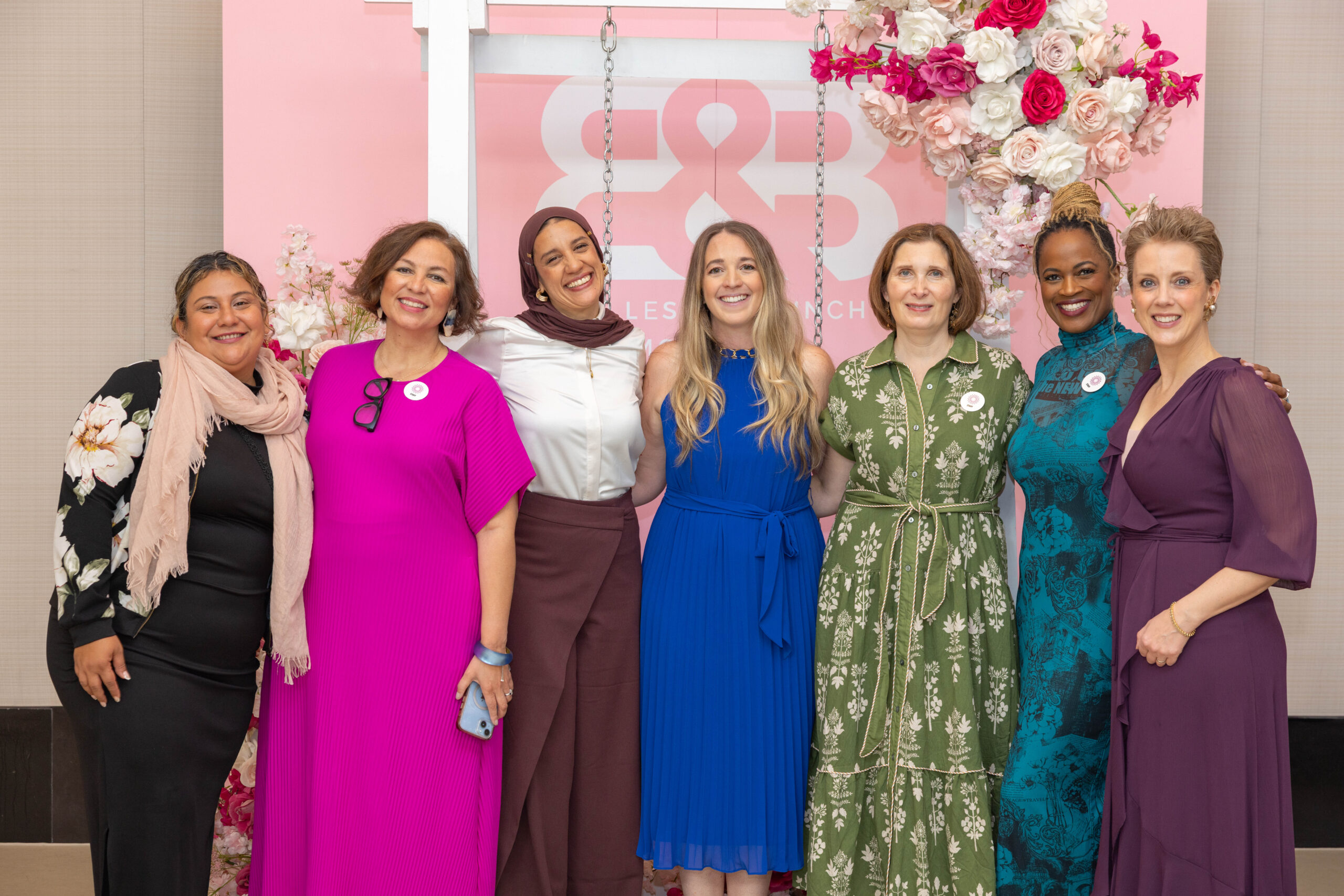 Seven smiling Look Good Feel Better staff pose together in front of a pink floral event backdrop, wearing colorful dresses and formal attire.