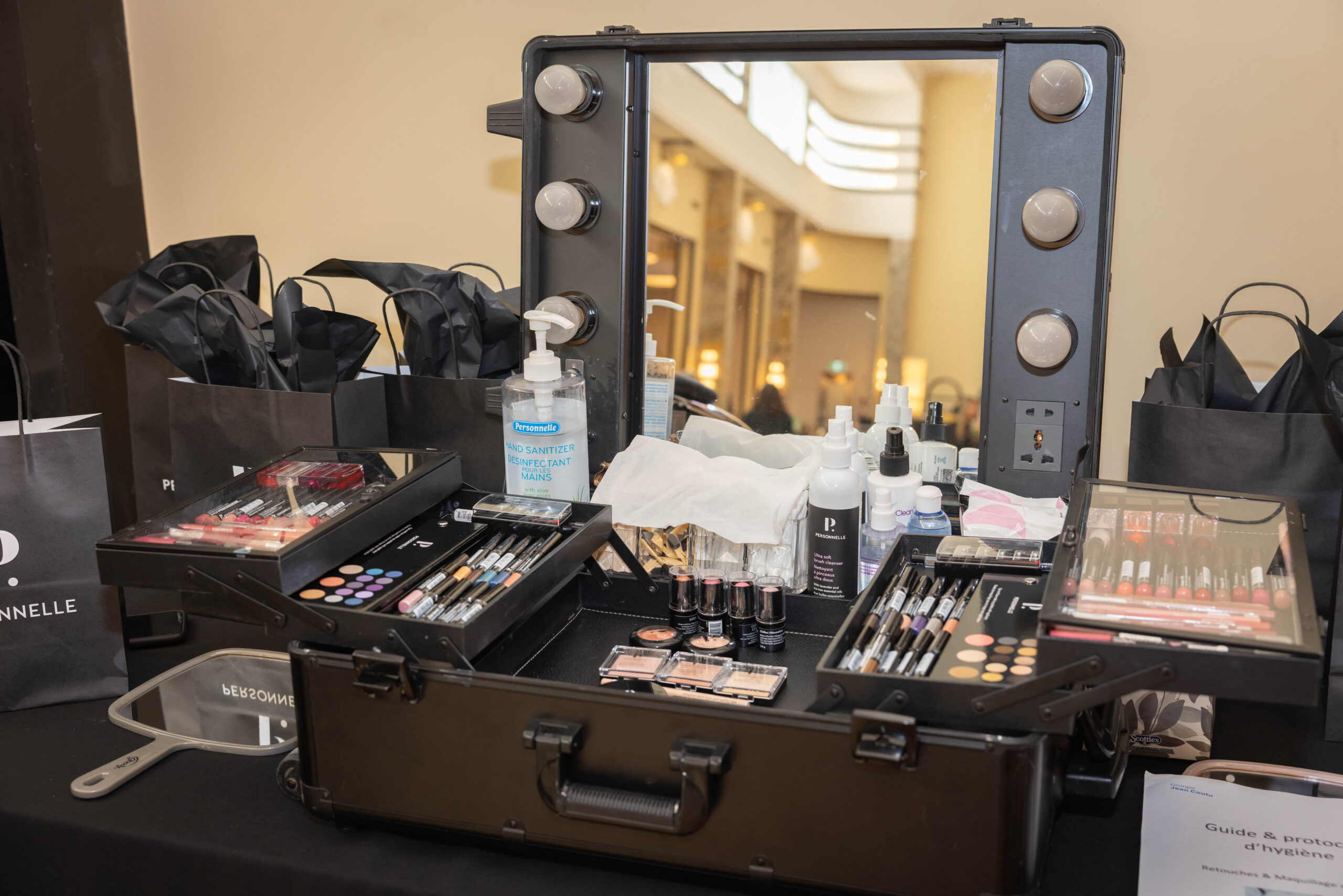 A professional makeup artist's open case with lit vanity mirror, brushes, eyeshadows, and beauty products at an event.