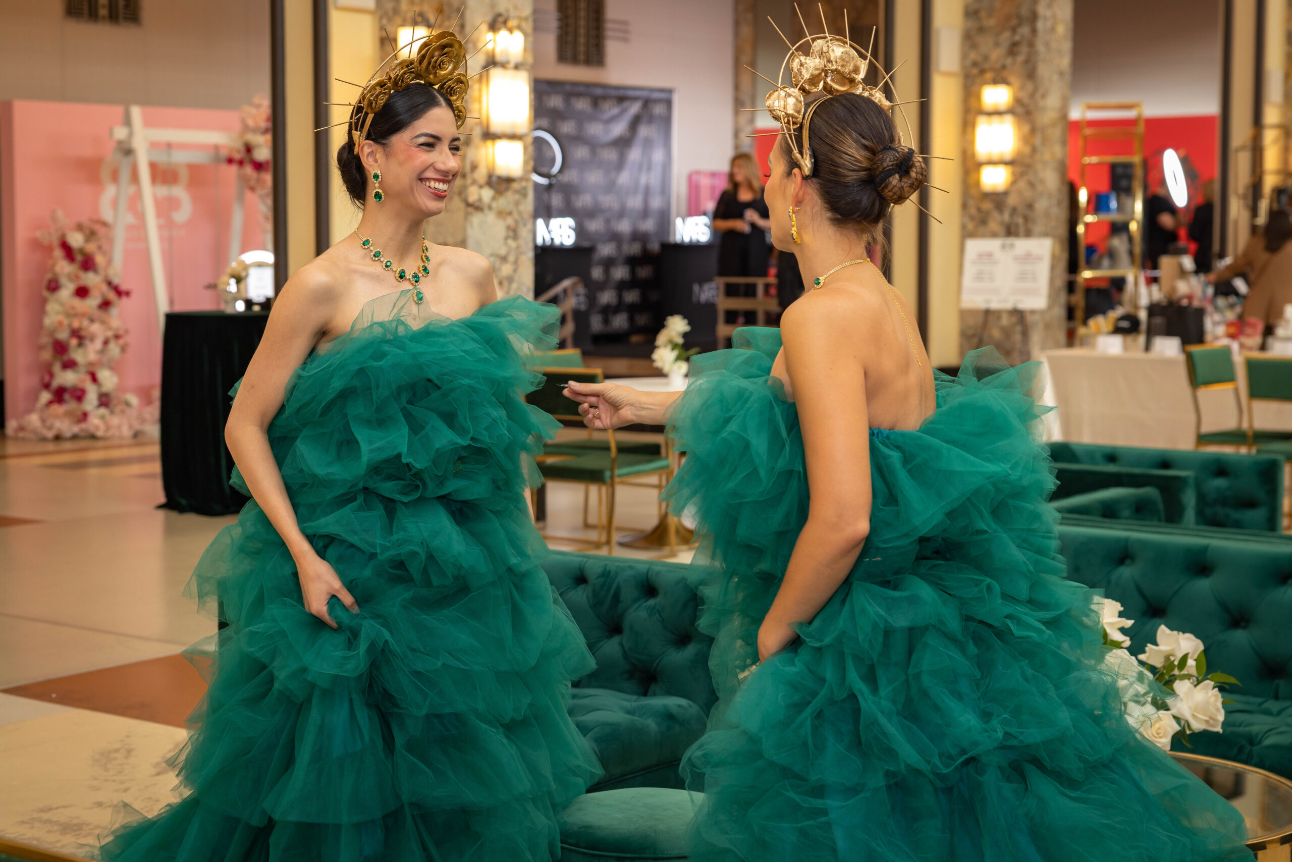 Two models in matching emerald green ruffled gowns and gold floral headpieces chat at an elegant indoor event.