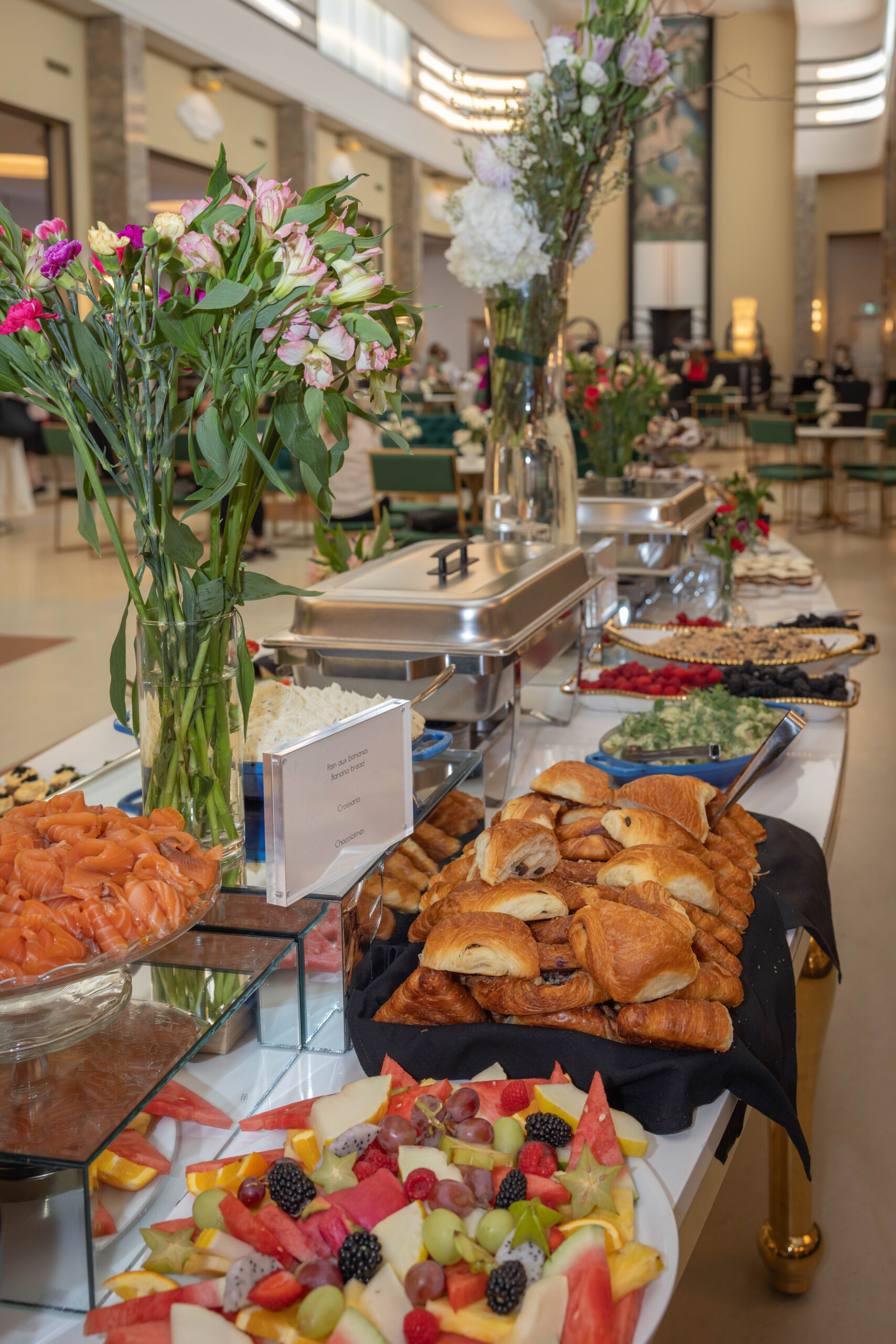 An elegant event buffet table with croissants, smoked salmon, fresh fruit, and floral arrangements in a grand hall.