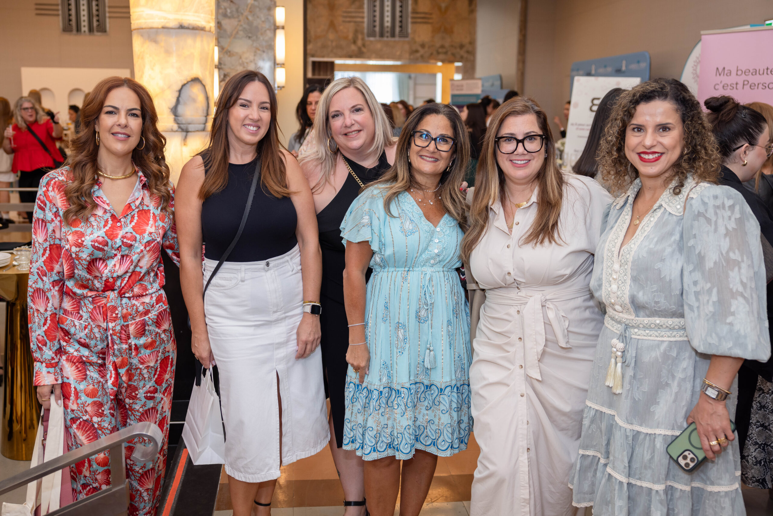 Six smiling women pose together at a busy indoor event, dressed in colorful and light-toned summer outfits.