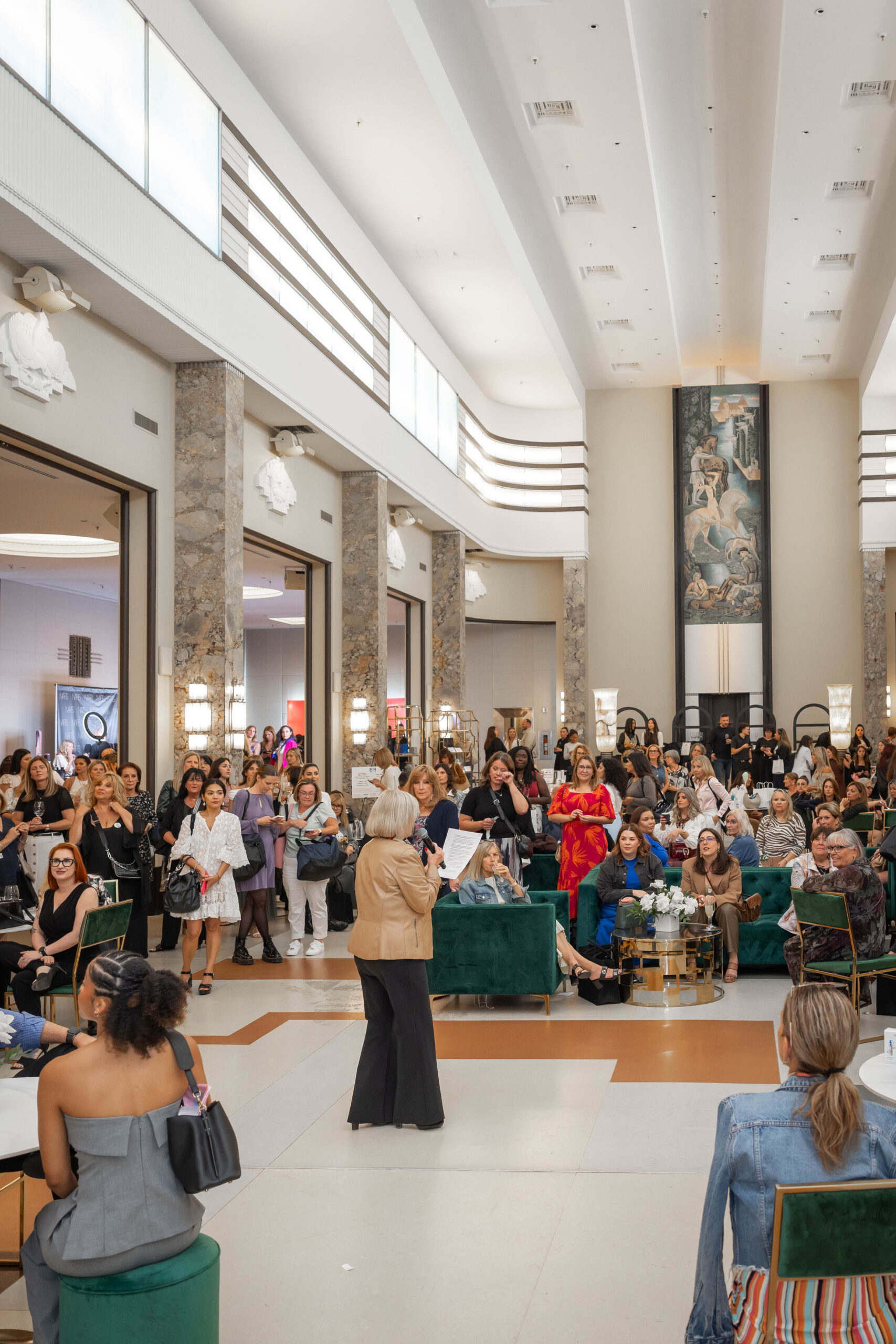 A speaker addresses a large crowd of women seated in a grand, high-ceilinged event space with green velvet furniture.