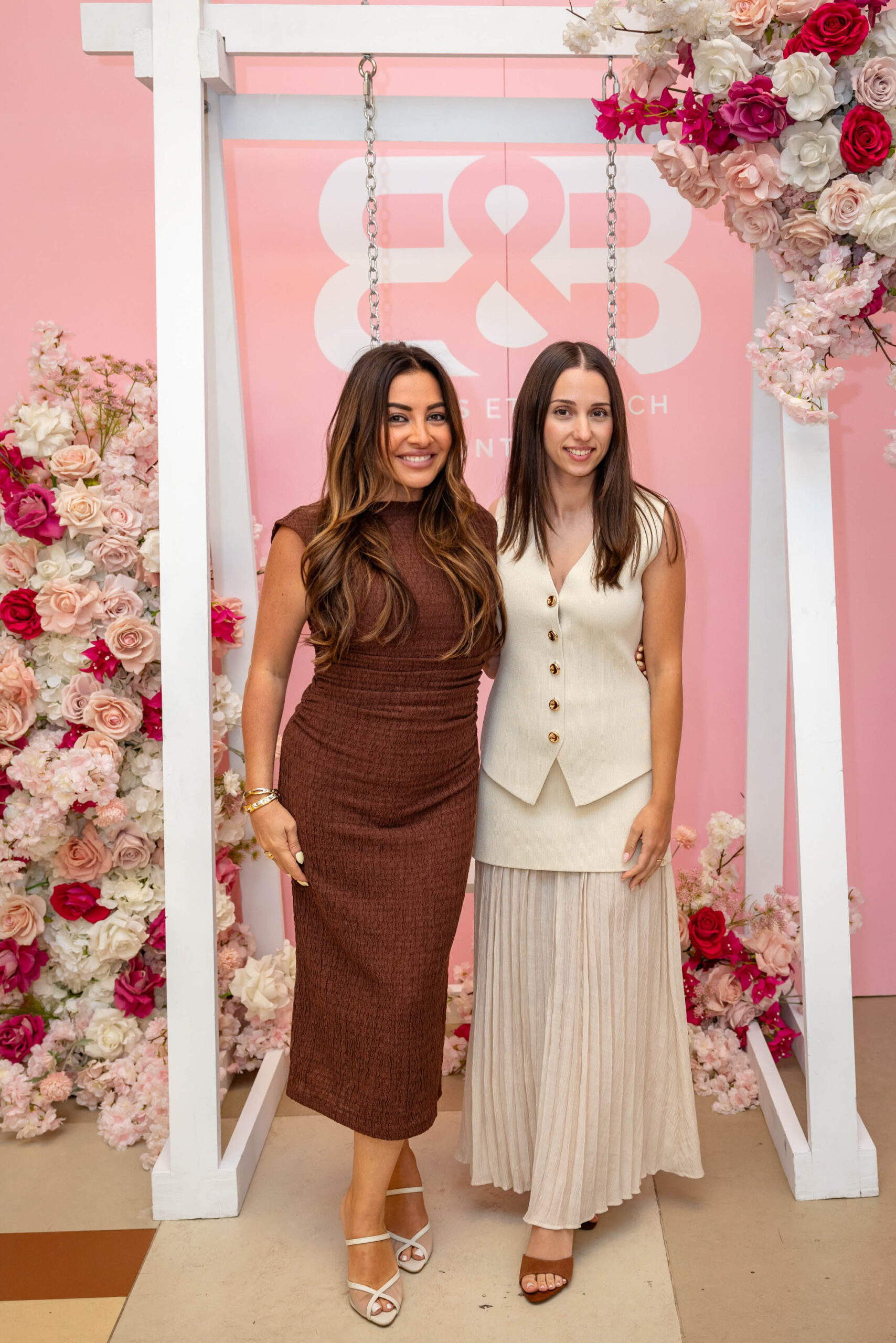 Two women pose together at a floral event display with a pink branded backdrop featuring roses and a white swing frame.