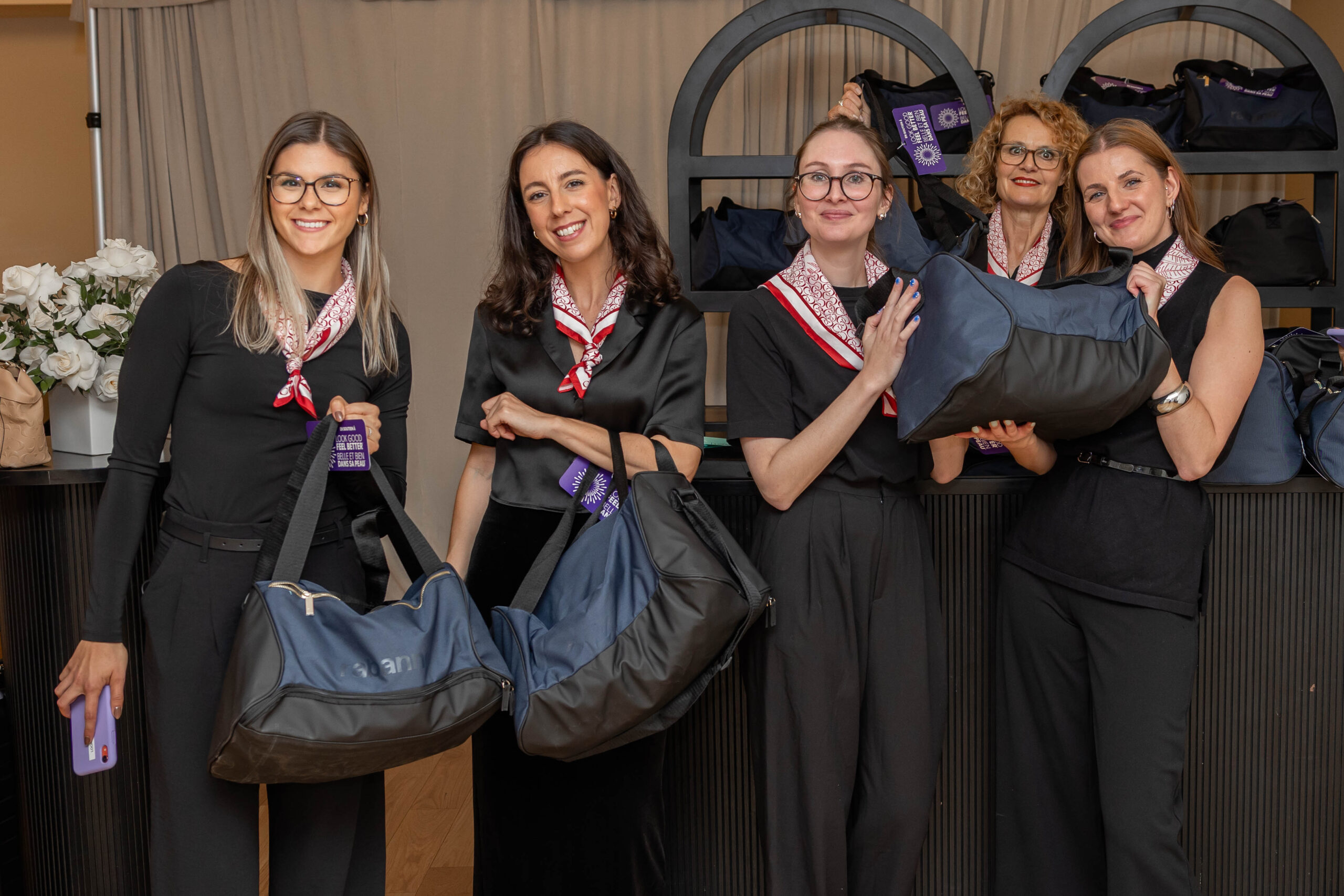 Five uniformed event staff in black outfits and patterned scarves smile while holding branded duffel swag bags.