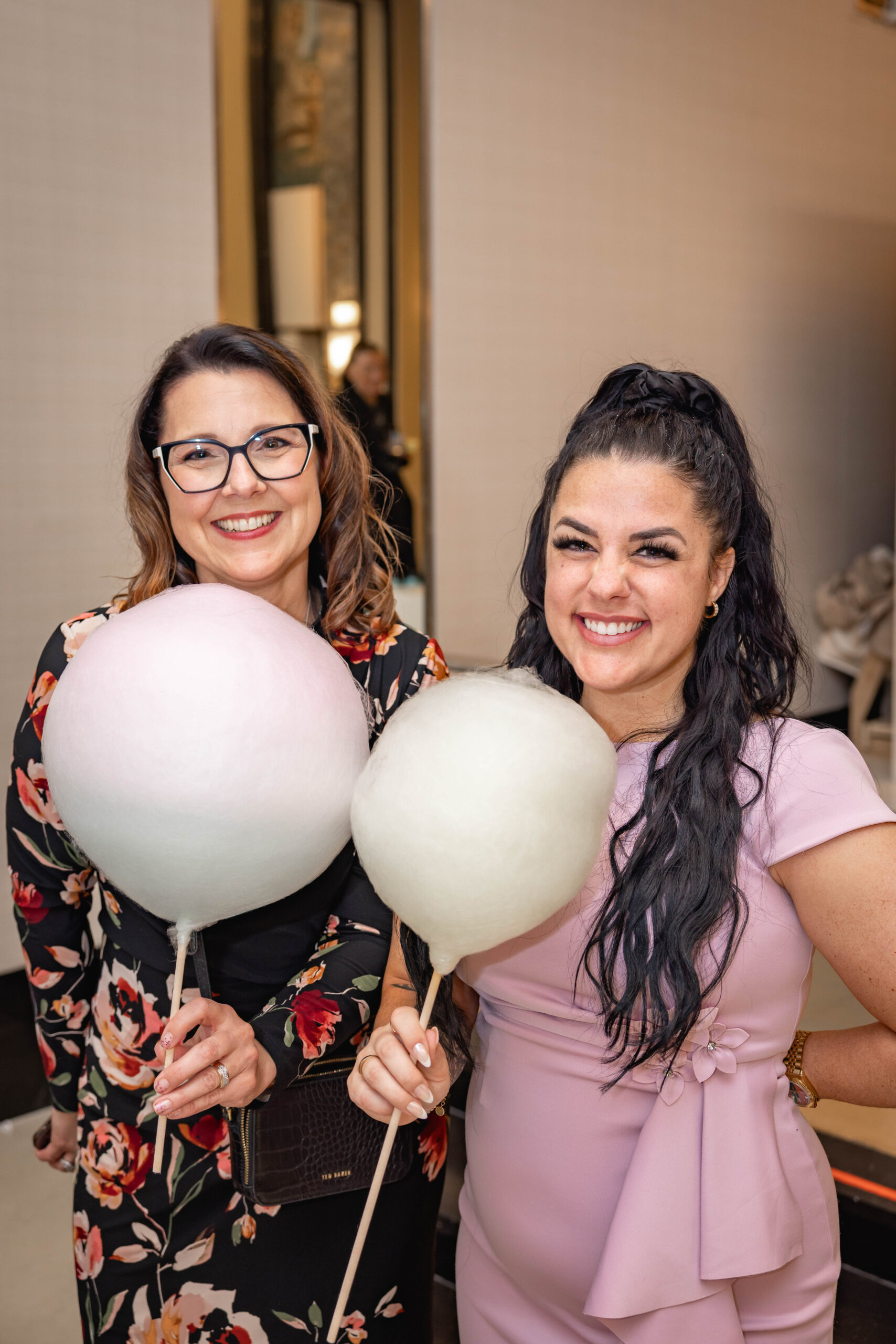 Two smiling women hold large cotton candy swirls on sticks at an event, one in a floral dress, the other in lilac.