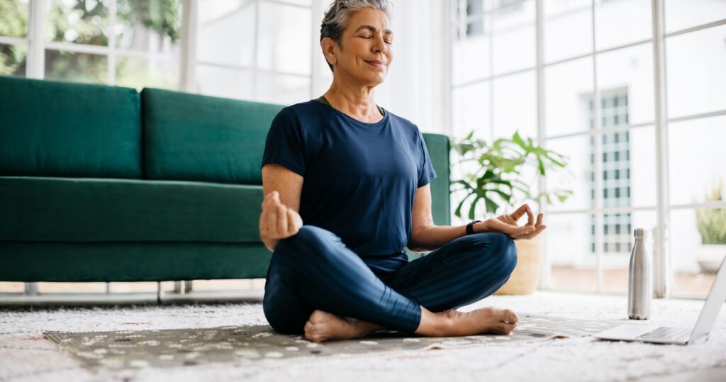 A woman meditating on a yoga mat in her living room, next to a laptop on the floor.